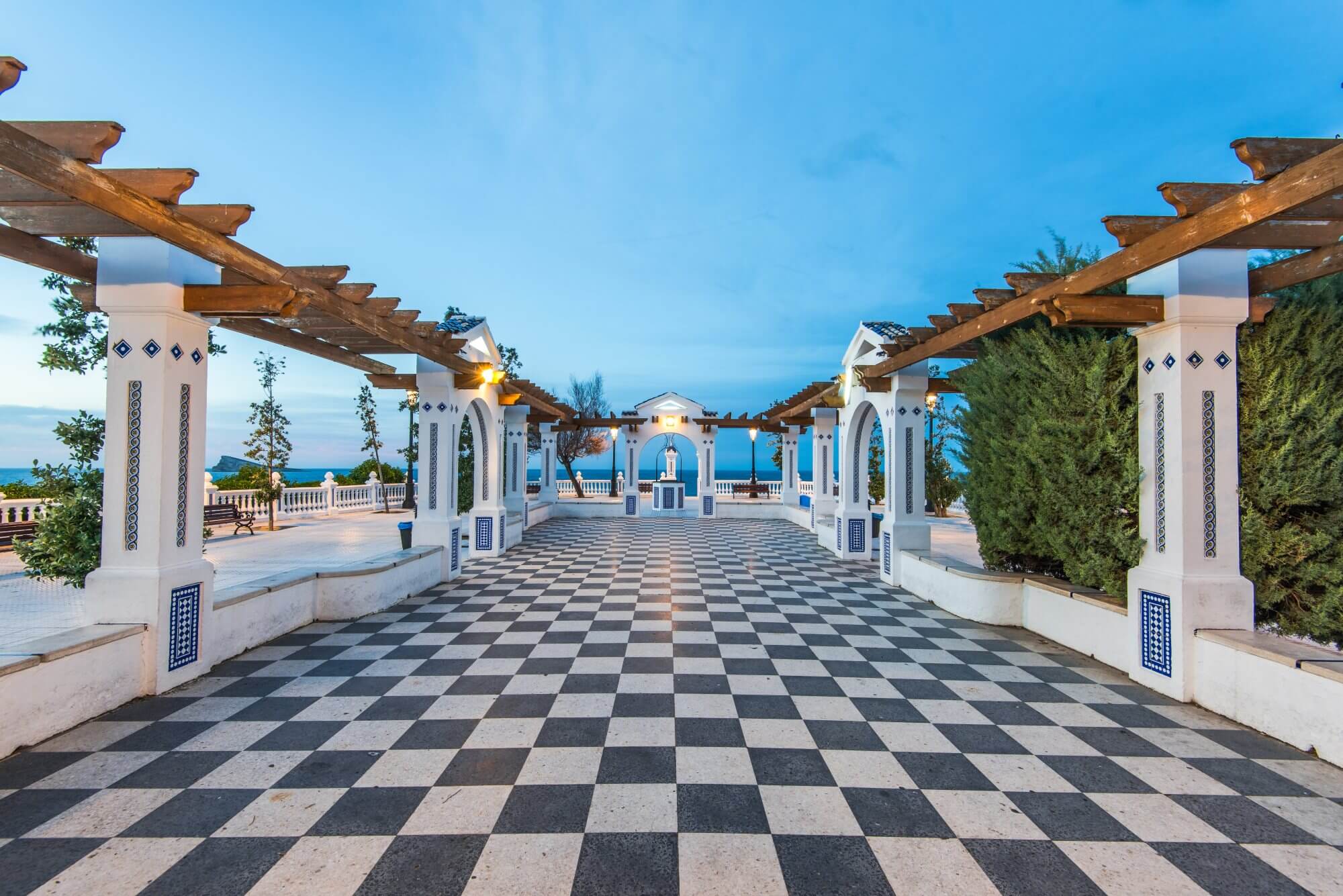 A black and white checked walkway framed by decorative white columns and landscaped plants leading to the sea