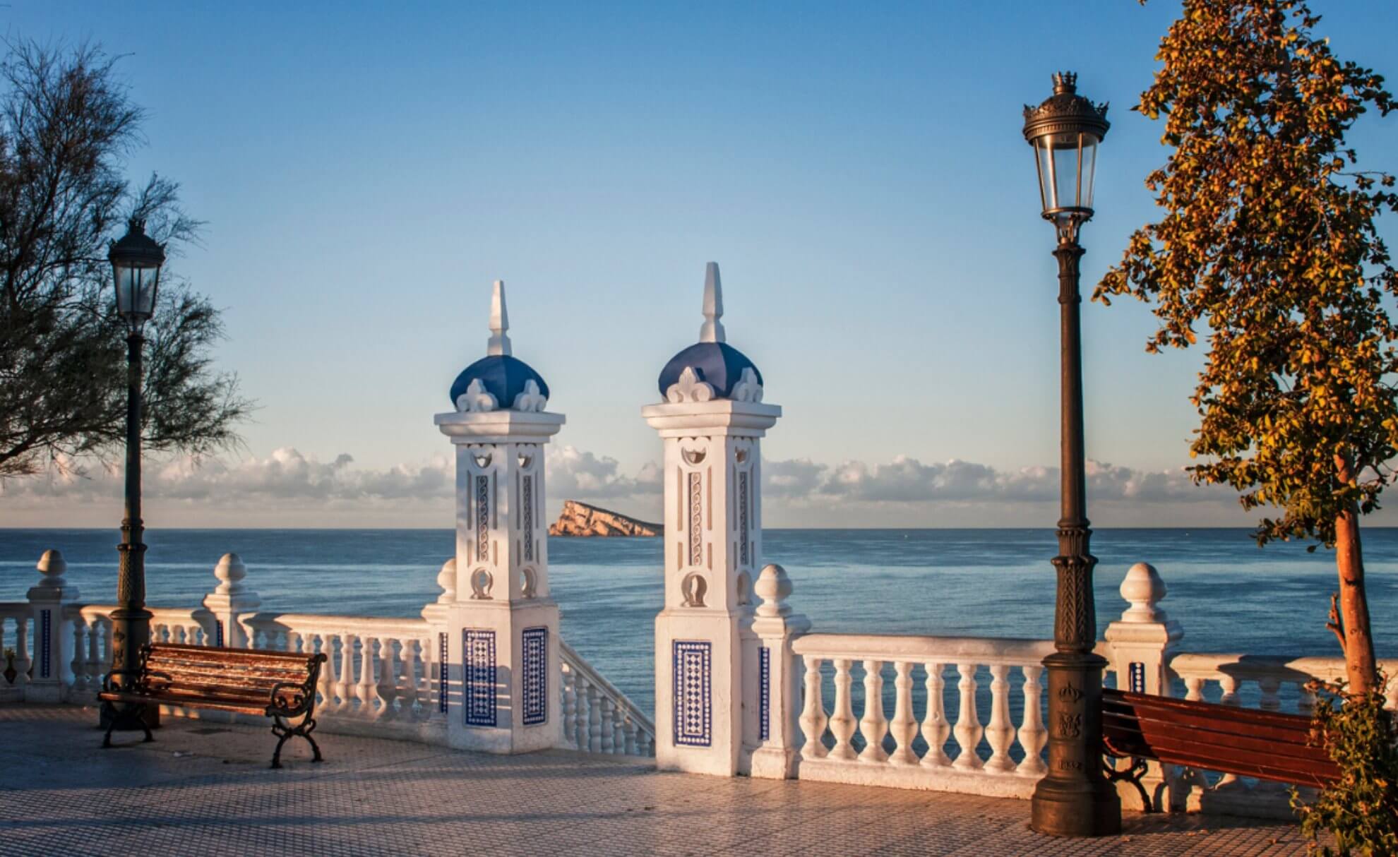 A paved path along the sea with lanterns, benches and white decorative columns